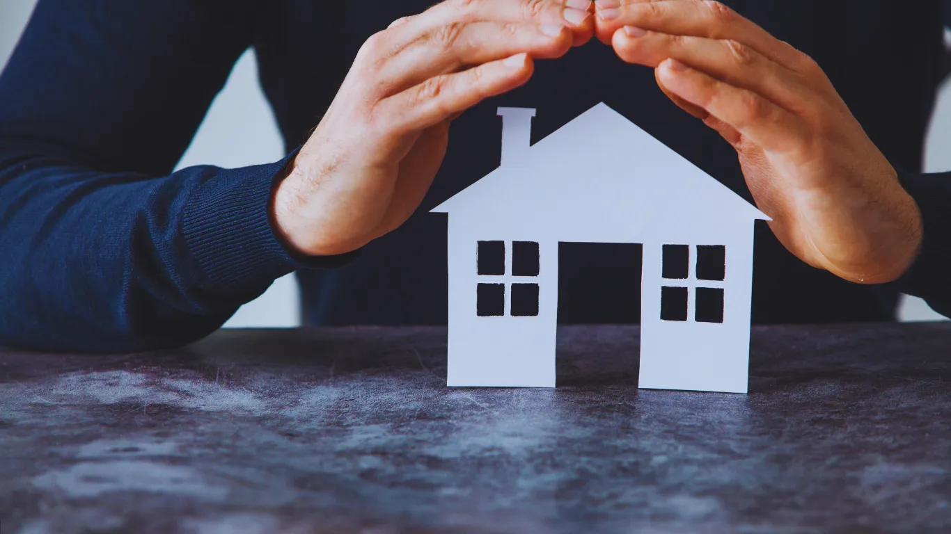 Hands protecting a paper house model, symbolizing home safety and roofing protection services.